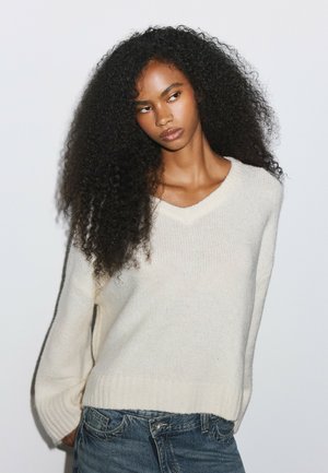 Femme aux longs cheveux bouclés portant un pull blanc en V et un jean bleu, debout devant un fond clair uni.