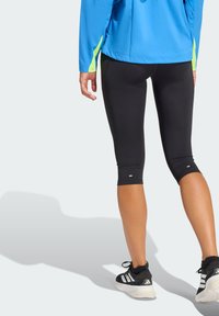 Mustat capri-leggingsit joustavasta kankaasta, joissa on sileä pinta ja polven pituinen leikkaus. Yhdistetty kirkkaan siniseen takkiin ja lenkkareihin.