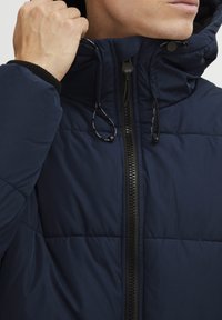 Námornícka puffer bunda s vysokým golierom, zipsom a nastaviteľnými šnúrkami. Tkanina s textúrou a prešívanými detailmi. Čierne akcenty.