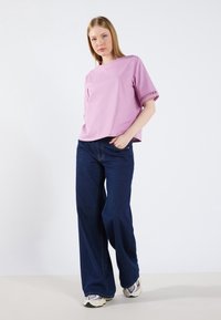 Rosa kortärmad topp med rund halsringning, ihop med lösa blå jeans och sneakers. Noterbara utskärningar vid ärmsluten.