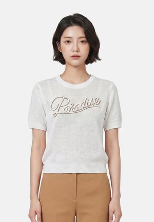Giovane donna con capelli corti e neri che indossa un top bianco a maglia a maniche corte con testo "Paradise" e pantaloni marrone chiaro, rivolta verso davanti.