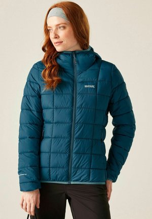 Kobieta o czerwonych włosach, ubrana w turkusową pikowaną kurtkę i szarą opaskę na głowę, patrząca w bok, trzymająca w lewej ręce czarny pasek.