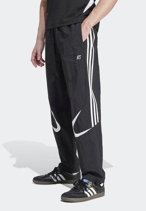 Osoba má na sobě černé tepláky s bílými pruhy a logem NTS, v kombinaci s černými teniskami Adidas s bílými pruhy.