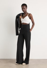 Schwarze weite Hosen kombiniert mit einem weißen, kurz geschnittenen Strickoberteil und einer schwarzen Lederjacke. Glattes Material mit subtiler Textur und metallenen Knopfdetails.