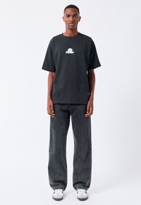 T-shirt in cotone nero con scollatura rotonda e maniche corte, caratterizzato da un logo grafico blu sul petto. Indossato con jeans scuri a vestibilità ampia.