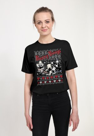 Jonge vrouw draagt een zwart Betty Boop T-shirt met cartoonfiguren, hartjes en sneeuwvlokpatronen op een witte achtergrond.