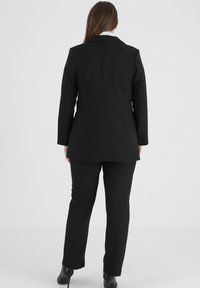 Den sorte dragt består af en skræddersyet blazer og lige bukser. Den har en glat tekstur, struktureret design og synlige sømme.