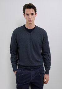 Pulover cu gât în V de culoare gri închis, realizat dintr-un material tricotat moale, cu mâneci lungi și croială lejeră, asortat cu pantaloni din catifea cu buzunare de culoare bleumarin închis.