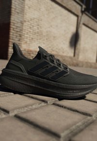 Черни маратонки Adidas Ultraboost с лека Boost подметка, поставени на каменно настилане на открито до тухлена стена.