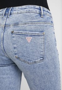 Jasnoniebieskie dżinsy z dopasowanym krojem, z postrzępionymi brzegami kieszeni oraz małym, trójkątnym logo "Guess" w kolorze czerwono-białym na tylnej kieszeni.
