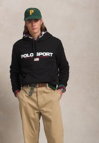 Črn kapuco s sweatshirtom z logotipom "POLO SPORT", khaki chinos hlačami in večbarvno karo srajco spodaj. Zeleni klobuk z rumeno črko P.