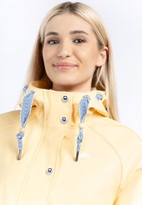 Gul vattentät jacka med hög krage, som har blå och vita randiga dragsnören, tryckknappar och en liten vit paraplylogotyp.