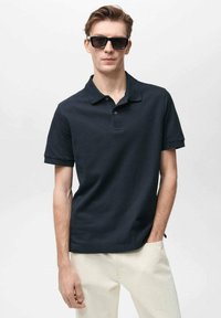 Námořnický polo s krátkým rukávem, zapínání na dva knoflíky a texturovanou tkaninou. Na sobě má světlé kalhoty a tmavé sluneční brýle.