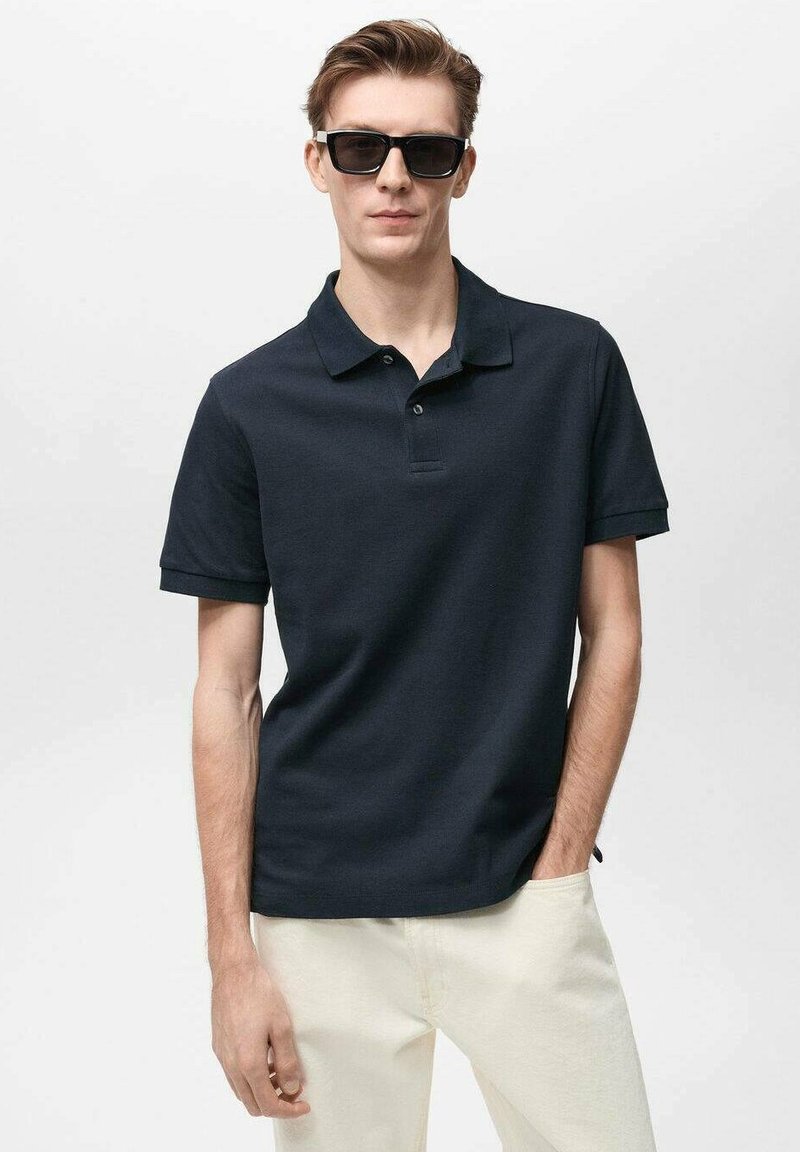 Námořnický polo s krátkým rukávem, zapínání na dva knoflíky a texturovanou tkaninou. Na sobě má světlé kalhoty a tmavé sluneční brýle.