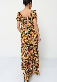 Robe maxi présentant un imprimé paisley noir, rouge et jaune, des manches courtes bouffantes, un dos carré et un corsage ajusté avec une taille froncée.
