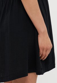 Rochie din material texturat negru care ajunge până la mijlocul coapsei, cu talie gathered și o croială lejeră. O mână este vizibilă ținând marginea fustei.