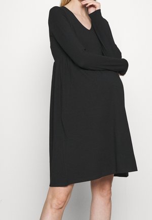 Femme portant une robe noire à manches longues jusqu'aux genoux avec un col en V, debout, une main près du menton et l'autre croisée à la taille.