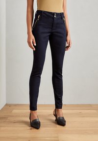 Jeans skinny de mezclilla en color náutico con cinturilla alta, que cuentan con dos bolsillos con cremallera dorada y cierre de botón, combinados con tacones negros de punta.