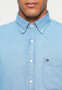 Ανοιχτό μπλε πουκάμισο από τζιν ύφασμα, με τσέπη που διαθέτει ένα μικρό λογότυπο, κλειστό γιακά και λευκά, μαργαριταρένια κουμπιά.