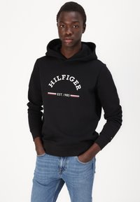 Sort hættetrøje med logoet "HILFIGER EST. 1985" i hvidt med røde detaljer, parret med blå jeans. Materialet ser blødt og tekstureret ud.