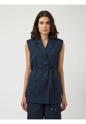 Kobieta o ciemnych włosach do ramion, ubrana w bezrękawnikowy granatowy marynarkę w prążki z podwójnymi guzikami oraz pasujące szerokie spodnie.