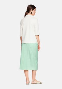 Weißes Kurzarmoberteil mit rüschigem Kragen, kombiniert mit einem mintgrünen plissierten Midirock. Das Model trägt beigefarbene Ballerinas.