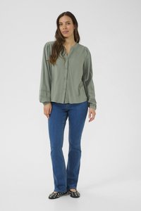 Camicia blusante verde chiaro con colletto rotondo, maniche arricciate e una sottile trama, abbinata a jeans blu aderenti e ballerine nere con borchie.