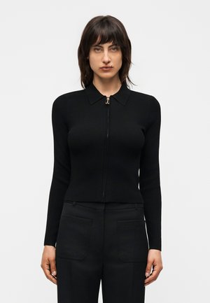 Sötét hajú nő, aki egy testhezálló fekete, cipzáras pulóvert gallérral és fekete, elülső zsebekkel ellátott nadrágot visel, egyszerű háttér előtt áll.