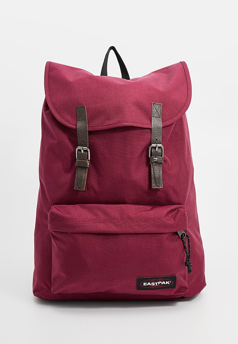 Sac à dos bordeaux avec un rabat en haut, deux bretelles marron et une poche zippée à l'avant. Fabriqué en tissu durable ; il possède un écusson logo et une boucle de suspendre.