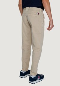 Pantaloni chinos beige in cotone con design aderente, tasche laterali e una tasca a toppa posteriore con un piccolo logo. Indossati con scarpe da ginnastica scure.