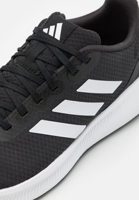 Scarpa da ginnastica nera con tessuto testurizzato, presenta tre strisce bianche sul lato, lacci neri e una soletta in gomma bianca.