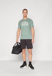 T-shirt vert menthe à manches courtes avec texte blanc, shorts noirs, baskets noires et un sac de sport noir. Style sportif simple.