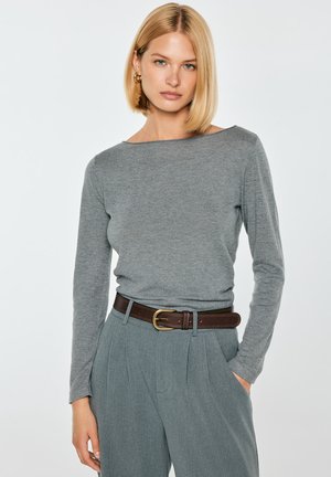 Kobieta z blond bobem, nosząca szarą bluzkę z długim rękawem, szare spodnie z wysokim stanem, brązowy pasek i złote kolczyki koła.