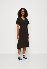Robe portefeuille noire à manches courtes, longueur genou, avec un accent à nouer à la taille, associée à des baskets montantes noires.