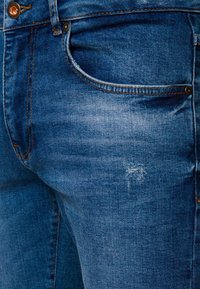 Modré džíny z denimu s mírně vybledlou texturou, designem s pěti kapsami a kontrastními měděnými akcenty. Výrazné třepení na okraji kapsy.