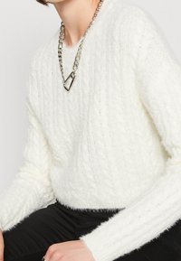 Vit kabelstickad tröja i mjukt, fluffigt material med långa ärmar och en kortare design, kombinerad med ett tjockt silverkedjehalsband.