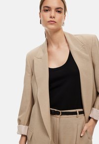 Beige bleiseri, joka on muotoiltu ja siinä on rakenteellinen muotoilu, varustettuna viistolla kauluksella ja raidallisilla hihasyistä, joka on yhdistetty mustaan toppiin ja kudottuun vyöhön.