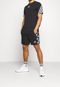 Hombre con pantalones cortos negros de Adidas con patrones laterales blancos, camiseta negra de Adidas con rayas blancas en las mangas, calcetines blancos de Adidas y zapatillas blancas.