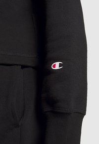 Sudadera negra con puños acanalados y un bolsillo canguro, con un pequeño logotipo de Champion en rojo y blanco en la manga izquierda.