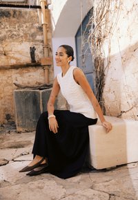 Femme tatouée assise sur un banc en pierre en plein air, portant un haut blanc sans manches, une jupe noire et des chaussures marron à bouts pointus, souriante et regardant sur le côté.