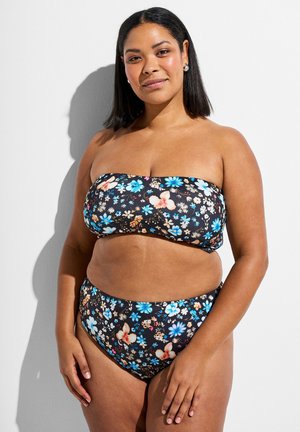 Kvindelig med kurver og skulderlangt hår iført sort blomsterbandeaubikini, stående mod en hvid væg med afslappede hænder.