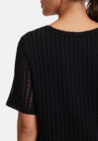 Schwarzes Strickoberteil mit einem durchbrochenen Design, runder Ausschnitt und kurzen Ärmeln. Der Stoff hat ein strukturiertes, atmungsaktives Aussehen.