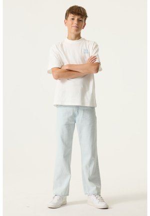 Witte T-shirt met korte mouwen en een klein blauw logo, gecombineerd met lichtblauwe jeans met rechte pijpen en witte sneakers. Eenvoudige, casual outfit.