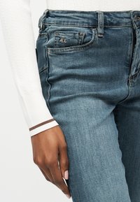 Κοντινό πλάνο χεριού ατόμου που ακουμπάει δίπλα στο σώμα, φοράει μπλε τζιν παντελόνι και λευκό πλεκτό μακρυμάνικο μπλουζάκι με καφέ ριγωτές μανσέτες.