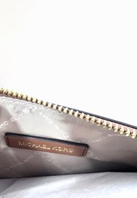 Brūnas ādas rāvējslēdzēja kabata ar zelta metāla rāvējslēdzēju, piedāvā sudraba logo oderi un zīmola nosaukumu, iecirstu zeltā.