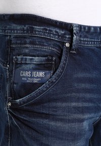 Donkerblauwe denimjeans met een versleten textuur, voorzien van een zichtbaar logo-label, een voorzak en metalen knoopdetails op de tailleband.