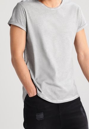 Mężczyzna w prostej, jasnoszarej koszulce z krótkim rękawem i podwiniętymi mankietami oraz czarnych, przetartych dżinsach, z rękami w kieszeniach, stojący na jasnym tle.