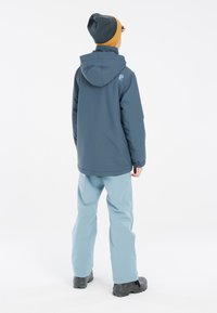 Giacca navy impermeabile con cappuccio, abbinata a pantaloni azzurri chiari e stivali neri. Presenta polsini elasticizzati e un dettaglio logo sulla schiena.