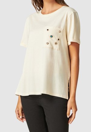 Frau trägt ein cremefarbenes kurzärmeliges Hemd mit einer Brusttasche, die mit kleinen, bunten Edelstein-Nieten verziert ist, kombiniert mit schwarzer Hose.