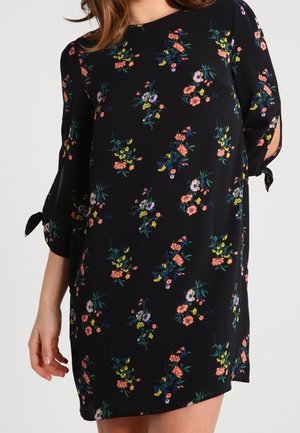 Robe noire à fleurs avec manches trois-quarts, poignets noués et col rond portée par une femme.